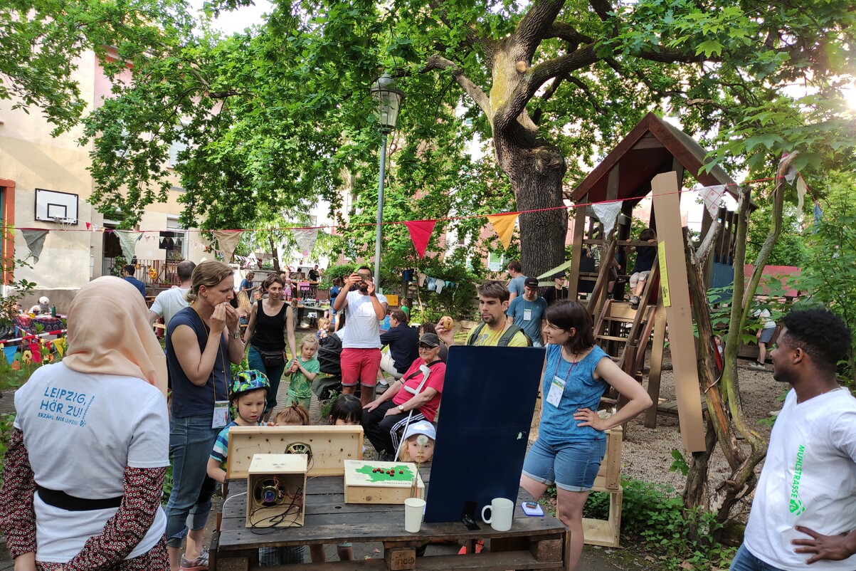 Viele menschen, eine Frau mit Kopftuch und einem T-Shirt mit dem Projektnamen sind zu sehen. Zentral sind Hör-Boxen, wie z.T. noch die Mikroelektronik zu sehen ist.